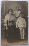   Retrato de Família / Maternidade / Genealogia Familiar Anterior. Retrato formal de uma mãe sentada em um banco de estúdio com seus dois filhos. À esquerda, a mãe exibe traje requintado de época com blusa de renda trabalhada. Ao fundo, uma menina em pé com vestido claro de punhos rendados e laço claro na lateral do cabelo. À direita, em primeiro plano, um menino posa com o clássico uniforme infantil de marinheiro (sailor suit) na cor branca, com gola e punhos listrados, calça comprida e sapatos escuros. Cenário de fundo difuso com coluna cenográfica. Características Físicas: Fotografia original em tom sépia, formato cartão-postal clássico com verso impresso em espanhol, corte reto tradicional.