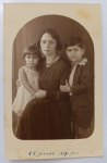   Retrato de Família / Maternidade / Linha Temporal Documentada. Composição em plano médio de uma mãe sentada ao centro, ladeada por seus dois filhos pequenos. À esquerda, uma menina com corte chanel veste um vestido claro leve e segura delicadamente o braço da mãe. À direita, um menino com expressão séria veste paletó escuro com colarinho de camisaria recortada em padrão de pontas claras por cima da lapela. Características Físicas: Fotografia original em tom sépia acentuado. Apresenta máscara fotográfica de estúdio em formato de arco ovalado (corte arqueado). Margem inferior com inscrição manuscrita a lápis: "12 anos depois". Corte reto tradicional.