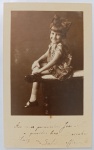  Retrato de Infância / Moda Infantil dos Anos 30 / Identificado. Retrato alegre e descontraído da menina Salvia sentada de lado sobre um móvel de estúdio (baú ou banco estofado), sorrindo para a câmera com uma perna cruzada. Ela veste um vestido estampado com motivos florais e babados, sapatos boneca escuros com meias claras de cano curto e ostenta um laço de fita agigantado no topo da cabeça, marca registrada da moda infantil de elite do período. Inscrições na Frente: Importante dedicatória manuscrita à tinta na margem inferior branca: "Aos meus priminhos José e Ilka e queridos tios Mariinha e Luiz - Salvia offerece." escrita com a grafia de época ("offerece").