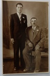  Retrato de Família / Paternidade / Imigração e Genealogia. Retrato formal de estúdio de pai e filho. À esquerda, o filho permanece em pé vestindo um elegante terno escuro acinturado, camisa de colarinho rígido, gravata listrada e uma flor (lapela/boutonnière) na jaqueta. À direita, o pai posa sentado em um banco de madeira, ostentando um bigode clássico, terno claro em tecido texturizado de abotoamento duplo, gravata listrada e flor idêntica na lapela. Cenário de estúdio com cortina escura ao fundo e piso de madeira. Inscrições no Verso: Rica dedicatória manuscrita à tinta, em idioma português, direcionada à destinatária Maria Rosa Estevas. Datada de "31-3-1941". Traz linhas guias impressas de cartão-postal ("Tarjeta Postal" esmaecido ao fundo) e marca d'água de fabricante alemã (Gevaert) na base.