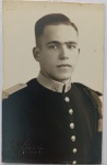  Retrato Militar / Militaria / Identificação Familiar. Retrato de estúdio em plano médio de busto de um jovem militar com expressão firme e serena. Veste túnica militar escura de abotoamento simples com botões metálicos abaulados reluzentes, ombreiras estruturadas (platinas) claras e gola alta rígida em tom claro com insígnias metálicas de arma ou unidade militar. Cabelo com corte regulamentar clássico de época. Características Físicas: Fotografia original em preto e branco com tom sépia suave, textura de emulsão fosca. Corte reto tradicional com bordas levemente rústicas.