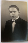  Retrato Masculino / Traje de Gala dos Anos 40 / Identificação Familiar. Retrato de estúdio em plano médio de busto de um jovem com expressão séria, identificado fisionomicamente como o mesmo militar do lote anterior de seu acervo. Veste um impecável traje formal composto por paletó escuro de lapela larga, camisa branca de colarinho alto com pontas viradas (gola borboleta) e uma elegante gravata borboleta escura. Cabelo curto cuidadosamente penteado para trás com fixador de época. Inscrições no Verso: Belíssima dedicatória manuscrita à tinta preta direcionada a "Titia e Dr. Heraclito", datada e localizada em Maceió. Linha divisória com a marca de papel postal nacional "WESSEL". O verso apresenta três grandes manchas escuras texturizadas de resíduo de cola e papel de álbum antigo, onde a foto esteve fixada.