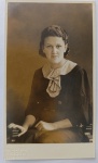   Retrato Feminino / Moda dos Anos 30 / Identificado. Retrato de estúdio em plano médio de três quartos da jovem senhora Maria do Carmo, posando sentada em uma cadeira de espaldar trabalhado. Ela exibe trajes e adornos característicos da alta moda dos anos 1930, destacando a blusa ou vestido escuro com gola clara estruturada em babados, broche geométrico e anel no dedo anelar esquerdo. Cabelo curto ondulado de época. Inscrições no Verso: Rica dedicatória manuscrita à tinta azul, assinada pela retratada ("Maria do Carmo"), direcionada à sua grande amiga "Maria Augusta". Divisória central com a marca de papel postal "WESSEL". Canto superior direito com um discreto carimbo em relevo invertido de fabricação do papel.