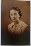  Retrato Feminino / Educação e Magistério / Moda dos Anos 30 / Identificado. Retrato de estúdio em plano médio de busto da jovem Hermé. Ela posa com expressão serena, ostentando trajes e corte de cabelo típicos da moda jovem da década de 1930, com blusa de gola geométrica detalhada e tiara fina nos cabelos cacheados. Inscrições no Verso: Rica dedicatória manuscrita à tinta, assinada pela retratada ("Hermé"), direcionada à sua professora "D. Maria Augusta". Verso limpo no formato clássico de cartão-postal, com um pequeno símbolo gráfico timbrado no topo da linha divisória central.