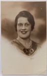  Retrato Feminino / Moda dos Anos 20/30 / Identificação Familiar / Possível Retrato de Dona Maria Augusta. Retrato de estúdio em plano médio de busto de uma senhora sorridente, exibindo trajes e adornos clássicos da transição da era das melindrosas para os anos 1930, destacando o colar de pérolas, penteado esculpido e flor de lapela (corsage). Características Físicas: Fotografia original em tom sépia médio de alta qualidade e nitidez de estúdio. Formato cartão-postal clássico com corte reto tradicional.