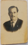 Retrato Masculino / Moda dos Anos 40 / Alfaiataria Risca-de-Giz / Identificação Familiar. Retrato de estúdio em plano médio de busto de três quartos do senhor Angelo na maturidade, personagem recorrente no arquivo de Dona Maria Augusta. Ele exibe trajes de alta costura masculina da época, destacando o terno risca-de-giz com pin de lapela e o penteado clássico com goma.