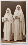  Retrato Religioso / Primeira Comunhão Coletiva / Arte Sacra / Identificação Familiar. Retrato formal de estúdio de duas meninas em trajes de comunhão solene de alta gala católica, com hábitos brancos, véus, terços, missais e arranjos florais.Uma análise fisionômica atenta revela traços familiares marcantes: a menina mais alta, à direita, compartilha uma semelhança física impressionante com a jovem Carmen (Bahia, Lote 32) e com a linhagem de Odette Oliveira Develly, apresentando o mesmo desenho de olhos expressivos e formato do rosto