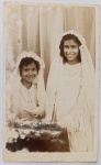  Tema: Retrato Religioso / Primeira Comunhão Coletiva / Infância / Identificação Familiar. Composição formal e alegre de duas meninas sorridentes em trajes de comunhão católicos, usando vestidos claros, véus longos e grinaldas florais. Ambientação de estúdio com cortina clara drapeada ao fundo. Características Físicas: Fotografia original em tom sépia/castanho de boa definição. Apresenta elegantes bordas totalmente decoradas em padrão serrilhado (deckle edge) nos quatro lados. Formato vertical.