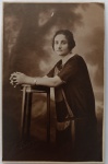 Retrato Feminino / Alta Sociedade Paulistana / Moda dos Anos 20 / Identificação Familiar. Retrato de estúdio em plano americano de uma senhora elegante apoiada em móvel de estúdio. Cenário de fundo pictórico pintado simulando um jardim ou bosque difuso.A estética da retratada é impecável e carrega o rigor da alta sociedade dos anos 1920: ela exibe cabelos escuros curtos penteados com coque baixo ou presos rente à nuca, lábios e sobrancelhas suavemente delineados e um relógio de pulso redondo com pulseira fina metálica (artigo de luxo e modernidade na época). Ela veste um suntuoso túnica ou vestido escuro em tecido leve com corte reto geométrico e painéis verticais contrastantes na parte frontal, típico da silhueta flapper. Ela posa em pé, de perfil, com os braços apoiados sobre uma banqueta alta de estúdio de madeira trabalhada e as mãos elegantemente postas com dedos entrelaçados.