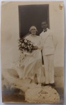  Retrato de Casamento / Moda Nupcial dos Anos 20 / Genealogia e Origem Familiar. Retrato formal de casamento em ambiente externo (provavelmente na saída da igreja ou residência), destacando a noiva com touca estilo melindrosa e buquê monumental, e o noivo com terno de gala branco e gravata borboleta. Características Físicas: Fotografia original em tom sépia médio de excelente definição. Formato cartão-postal clássico com corte reto tradicional.