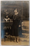 A indumentária e os adornos da retratada refletem com máxima fidelidade o auge da moda burguesa urbana dos anos 1920. O grande destaque é o longo colar de duas voltas de pérolas, joia emblemática do figurino das "melindrosas". Ela veste um suntuoso vestido escuro de cintura baixa, com corpete liso e saia fluida levemente rodada, complementado por mangas e peitilho em renda fina trabalhada. Nos pés, ostenta sapatos bicolores (claro e escuro) com fivela e salto baixo de época. O cabelo curto exibe o penteado característico com mechas moldadas fixas na testa e têmporas.A ambientação é cênica e realista, realizada na área externa de uma residência ou estúdio com escadaria de pedra, coluna de madeira e uma imponente grade de ferro fundido ornamental ao fundo. À esquerda, há um pedestal alto de madeira sustentando um vaso com flores (antúrio) sobre uma toalha rendada, e uma pequena palmeira na prateleira inferior.