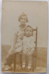  Retrato de Infância / Irmãos / Moda Infantil de Época / Identificação Familiar. Retrato vernacular de duas crianças sentadas em uma cadeira de madeira na área externa de uma residência (junto a uma parede texturizada), exibindo trajes, calçados e cortes de cabelo típicos das décadas de 1920 e 1930. Características Físicas: Fotografia original em tom sépia/castanho de boa definição e pátina suave do tempo. Formato cartão-postal clássico com corte reto tradicional.