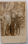   Retrato Masculino / Alta Moda dos Anos 20 / Chapéu Boater / Identificação Familiar. Retrato formal em plano inteiro de três jovens cavalheiros posando de braços dados em um parque ou jardim residencial, exibindo ternos completos e os tradicionais chapéus de palha canotier. Características Físicas: Fotografia original em tom sépia/castanho de excelente definição e nitidez, com rica pátina do tempo. Formato cartão-postal clássico com corte reto tradicional e margem branca inferior para dedicatória.