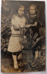  Retrato de Infância / Moda dos Anos 20 / Elemento Cotidiano / Identificação Familiar. Retrato vernacular em plano inteiro de duas meninas posando de braços dados em um jardim residencial atrás de um vaso improvisado de samambaia em lata litografada antiga de "10 Kilos". Características Físicas: Fotografia original em tom sépia/castanho de boa definição de primeiro plano. Formato cartão-postal clássico com corte reto tradicional. Vertical.