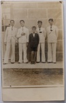  Fotografia Antiga / Cartão-Postal Fotográfico Vernacular de Época (Real Photo Postcard - RPPC). Época Estimada: Circa 19321938. Procedência: Provável Maceió ou Rio de Janeiro (Eixo Família Oliveira Develly). Tema: Retrato de Família / Irmãos / Alfaiataria Antiga / Rito de Luto / Identificado. Retrato formal em plano inteiro de cinco irmãos posando alinhados, destacando quatro rapazes em ternos trespassados brancos de linho (dois com tarjas de luto na lapela) flanqueando o irmão menor ao centro em traje escuro tradicional com bermuda.