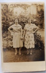  Fotografia Antiga / Cartão-Postal Fotográfico Vernacular (Real Photo Postcard - RPPC). Época Estimada: Circa 19401948 (Meados da década de 1940). Tema: Retrato Feminino / Moda dos Anos 40 / Lazer Urbano / Identificação Familiar / Círculo de Maria Augusta. Retrato de corpo inteiro de duas senhoras de mãos dadas em um parque ou jardim público, exibindo vestidos e conjuntos de alta costura civil de passeio e calçados da época, com arquitetura modernista ao fundo.