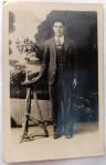   Fotografia Antiga / Cartão-Postal Fotográfico de Estúdio (Real Photo Postcard - RPPC). Época Estimada: Circa 19201928 (Anos Loucos). Procedência Histórica: Provável Maceió ou Rio de Janeiro (Conexão direta com o Eixo Família Oliveira Develly). Tema: Retrato Masculino / Alta Moda dos Anos 20 / Alfaiataria Xadrez / Iconografia de Elite / Genealogia Familiar Anterior. Retrato formal em plano inteiro de um jovem cavalheiro posando em pé em estúdio ao lado de um pedestal com arranjo de flores, exibindo terno xadrez completo de três peças (com colete) de alta alfaiataria e cenário pictórico de paisagem ao fundo.