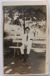  Fotografia Antiga / Cartão-Postal Fotográfico Vernacular de Época (Real Photo Postcard - RPPC). Época Estimada: Circa 19261934. Procedência Histórica: Provável Maceió ou Rio de Janeiro (Conexão direta com o Eixo Família Oliveira Develly). Tema: Retrato Masculino / Moda dos Anos 20 e 30 / Rito de Luto Tradicional / Braçadeira de Luto / Identificação Familiar. Retrato formal em plano inteiro de um jovem cavalheiro sentado em um banco de praça pública, vestindo terno completo branco com braçadeira de luto preta na manga esquerda, exibindo os costumes de respeito e etiqueta civil de época.
