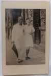   Fotografia Antiga / Fotografia de Rua Vintage (Street Snapshot / Lambe-Lambe). Época Estimada: Circa 19401948. Tema: Vida Urbana / Cotidiano / Moda dos Anos 40 5 / História da Fotografia / Identificação Familiar. Registro instantâneo de calçada mostrando o patriarca sênior da família de terno branco e gravata borboleta caminhando de braços dados com uma jovem senhora em frente a um comércio antigo com a placa "FOTOGRAFIAS AQUI".