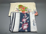 CAMA - MESA - BANHO - Lote de duas toalhas de mesa em algodão (200 cm x 120 cm cada). A primeira possui fundo creme com arranjo floral em tons de rosa, verde e azul, com acabamento em viés azul marinho. A segunda traz faixas verticais e barrado central em fundo azul marinho com flores em tons de vinho e rosa. Marcas do tempo e de uso.