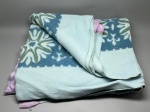 CAMA - MESA - BANHO - Coberta de solteiro em tecido aveludado/lã em tons de azul e verde-água, decorada com faixa central com padronagem geométrica e floral estilizada. A peça possui acabamento nas bordas em fita de cetim na cor lilás e apresenta marcas do tempo, de uso e pequenos desgastes.