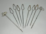 METAL - Conjunto com 8 espetos para aperitivos ou petiscos em metal espessurado a prata, sendo dois com ponteira decorativa em relevo representando cervo sobre folhagem e seis com cabo em formato de losango/olho com haste torcida. Apresentam desgastes no banho, oxidação e marcas do tempo. Alt. aproximada 20 cm.