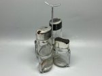 METAL E VIDRO - Galheteiro/porta-condimentos de mesa composto por suporte metálico cromado com alça central arqueada e quatro recipientes em vidro transparente com tampas dosadoras metálicas (sendo dois maiores e dois menores). As peças apresentam desgastes, manchas de oxidação, resíduos internos e marcas do tempo.