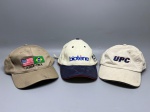 DIVERSOS - Lote com três bonés promocionais e corporativos variados, sendo um boné bege "ARM-TEX" com bordado frontal contendo as bandeiras dos Estados Unidos e do Brasil e marcas de uso; um boné "Biotène" em tom off-white com aba em camurça sintética azul-marinho, detalhe vermelho na borda e bordado frontal da marca junto a um bordado lateral alusivo a evento internacional; e um boné "UPC" em tecido algodão cru com aba estruturada, bordado frontal em destaque com a sigla em azul com detalhe em vermelho, contendo pequenos desgastes e marcas naturais do tempo.