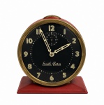 SMITHS ALARM  Antigo relógio despertador inglês, com caixa em acabamento vermelho, mostrador preto com numerais em tom marfim, aro em metal dourado e mecanismo mecânico. Peça marcada MADE IN GREAT BRITAIN, de estética vintage. Funcionamento desconhecido. Medidas: 11 x 10 cm.