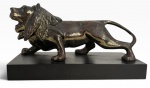Leão em bronze patinado, de expressiva modelagem naturalista, em posição de marcha, sobre base retangular em madeira. Apresenta pátina em tons castanho e dourado, com detalhes da juba e musculatura destacados. Peça de caráter decorativo e escultórico. No verso, etiqueta da ANTIGA METAIS ARTÍSTICOS LTDA.  São Paulo. Mede 19cm x 10cm.
