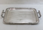 WOLFF PRATA 90. Elegante bandeja retangular confeccionada em metal espessurado à prata, centro ligeiramente rebaixado, borda contornada por fileira de frisos, flores e fitas em relevo, alças laterais fusiformes, cobertas por folhagens, apoiadas sobre hastes curvadas. Marca da manufatura no verso. Leve desgaste no prateado. Med.: Alt. 6 x Comp. 48 x Larg. 32,5 cm.