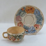 Xícara de  chá de coleção em faiança européia, decorada com flores e folhagens douradas em alto relevo, em rica policromia, pires com folhagens e farta douração. Marcada. Med. 7 x 10 cm.