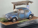 Miniatura die-cast do Volkswagen Fusca (1985) na cor azul, apresentada num diorama personalizado com efeito envelhecido e enferrujado (barn find), sob um telheiro rústico de madeira com cobertura de zinco, placa decorativa e base com vegetação e solo simulados. Medidas: 05 x 10 x 05 cm. Com marcas de tempo de uso.