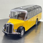 Miniatura colecionável em die-cast (metal com pormenores em plástico) na escala 1/43, referente ao autocarro clássico Saurer L4C. Apresenta pintura na cor amarela vibrante com teto e contornos de janelas prateados, além de guarda-lamas pretos. Destaca-se pelo rico detalhamento, incluindo grelha frontal vertical com o emblema "Saurer", faróis circulares dianteiros, insígnia da PostAuto/correios suíços na frente, para-brisas divididos com limpadores em miniatura, janelas superiores no teto com acabamento escurecido, cúpula superior ventilada, placa identificativa "P 23123" e interior visível com assentos e volante brancos. Equipado com jantes prateadas e pneus duplos no eixo traseiro. O chassis inferior rígido apresenta a gravação em relevo original "SAURER L4C 1/43"