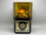 Relógio de mesa de movimento quartz composto por base octogonal em plástico preto com mostrador redondo de fundo dourado, ponteiros pretos e ponteiro de segundos vermelho, emoldurado por moldura metálica com acabamento dourado e motivos arabescados. Sobre a base, câmara hexagonal em acrílico âmbar com composição decorativa interna representando peixes e plantas aquáticas em fibras sintéticas nas cores dourado e laranja. Mostrador exibe inscrição "QUARTZ". Sinais de uso e oxidação na moldura metálica. Sem marca identificada. Funcionamento não verificado. Med. 37x20 cm.