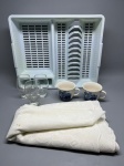 Lote composto por: escorredor de louças em plástico branco com suporte para pratos e talheres; três taças em vidro transparente com base pedestal e corpo tulipa, sem decoração; duas canecas em cerâmica branca com decoração em azul cobalto representando cena arquitetônica; e duas peças de tecido dobradas em tom off-white com textura jacquard. Itens apresentam sinais de uso. Não identificada marca ou procedência nos itens visíveis nas imagens. copo com pé, xícara grande, porta-louças, enxoval, conjunto de mesa, toalha