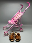 Lote composto por dois itens. Carrinho de boneca dobrável, estrutura tubular metálica na cor rosa claro, assento em tecido estampado com flores e inscrições em tom rosa escuro sobre fundo creme, quatro rodas duplas em plástico rosa e branco, guidão com empunhadura em plástico rosa escuro. Apresenta sinais de uso. Acompanha par de sandálias infantis em couro marrom, modelo aberto com tiras entrelaçadas e fivela lateral, solado em material claro. Estado de conservação compatível com uso. carrinho de bebê em miniatura, calçado infantil, pushchair de boneca, sandália de criança, toy stroller