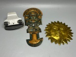 Lote composto por três peças. Furador de papel de mesa com base oval em plástico cinza e alavanca em plástico branco, mecanismo metálico exposto, com mola aparente; apresenta desgastes de uso. Placa decorativa em metal com acabamento em tons de verde e dourado, representando figura estilo pré-colombiano com ornamentos e tocado, com sinais de desgaste na pintura. Aplique decorativo em metal dourado em formato de sol com rosto antropomórfico em relevo e raios ao redor. Peças sem marca identificável. Estado de conservação variado. perfurador, mascara andina, inca, rosto solar, adorno, ornamento, artefato decorativo, sol antropomórfico