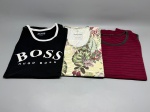Lote composto por três camisetas masculinas usadas. A primeira é uma regata preta com vivos brancos nas bordas, estampa frontal com logotipo 'BOSS Hugo Boss' em branco e etiqueta interna da marca Hugo Boss, informando 100% cotton. A segunda é uma camiseta de manga curta com estampa floral tropical em tons de verde, amarelo e rosa, com etiqueta da marca Mahalo. A terceira é uma camiseta de manga curta na cor vinho com listras horizontais em preto e acabamento contrastante na gola. Tamanhos não verificados individualmente. Estado de conservação: usadas. blusa, camisa, peça casual, indumentária, vestuário masculino, top, conjunto de roupas