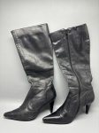 Par de botas femininas de cano longo, na cor preta, confeccionadas em material de aspecto liso e brilhante, aparentando couro. Bico fino pontiagudo, fechamento lateral por zíper ao longo do cano. Salto tipo agulha de média altura. O forro interno aparenta tecido escuro. As peças apresentam marcas de uso, com vincos e desgastes visíveis no cano e na biqueira. Numeração não identificada nas imagens. Sem marca visível nas fotografias. Lote composto por duas peças (par). botas de cano alto, bota de salto, calçado feminino, bota preta, bota zíper