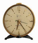 WESTCLOX  Relógio de mesa vintage, mostrador em tom marfim com numerais e ponteiros dourados protegidos por vidro de formato convexo, caixa em metal com pátina e marcas naturais do tempo, sobre suporte. Medidas: 14 x 14 cm.