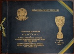 PEÇA DE MUSEU: Álbum Ano de Ouro do Esporte Brasileiro 1959 - AUTOGRAFADO por Thomaz Mazzoni - Relíquia da Copa de 58Descrição do Lote:Raridade documental absoluta, com valor de associação histórico e procedência inquestionável. Trata-se do álbum comemorativo oficial de luxo "Ano de Ouro do Esporte Brasileiro", publicado em 1959 em comemoração ao primeiro aniversário da conquista do inédito título mundial de futebol na Suécia (1958). Uma das publicações mais imponentes e raras da história da Seleção Brasileira, editada sob os auspícios da C.B.D., F.P.F. e do Dr. Paulo Machado de Carvalho (Chefe da Delegação). AUTÓGRAFO HISTÓRICO: A folha de guarda traz a assinatura autógrafa original e legítima de Thomaz Mazzoni (Olympicus), o genial diretor e idealizador da obra, tornando este exemplar uma peça única no mundo de valor iconográfico imensurável. Direção e Realização: Bureau Interamericano de Imprensa  Direção de Thomaz Mazzoni e Com. Lauro Luz. Procedência / Homenagem de Época: Capa personalizada na margem inferior com a inscrição dourada de homenagem do histórico clube do interior paulista E. C. São Bento de Marília. Riqueza Gráfica e Artística Extraordinária: Abre com uma belíssima e poética folha de rosto inteiramente desenhada e colorida no padrão aquarela de época, exibindo a bandeira nacional, o pavilhão sueco, o contorno cartográfico do Brasil e a Taça Jules Rimet. Apresenta crônicas monumentais escritas em tempo real sobre a constelação de campeões de 58, destacando o capítulo histórico de Pelé aos 17 anos (descrito por Mazzoni como o "L'Enfant Terrible, o menino que assombra o mundo" e detalhando sua recuperação milagrosa do joelho), as biografias de Mazzola, e páginas dedicadas aos arqueiros e suplentes como Moacyr e o goleiro Castilho.Estado de Conservação (Conforme Fotos): Capa (Encadernação de Luxo Almofadada): Original em percalina ou couro sintético texturizado azul-marinho com gravações em hot-stamping dourado (Brasão da República dos Estados Unidos do Brasil e a Taça Jules Rimet) em excelente estado de rigidez e conservação. Lombada firme amarrada com o cordão e franja original texturizada verde e amarela de época. Discretas marcas de armazenamento nas bordas externas. Miolo: Espetacular estado de conservação. Páginas com formato panorâmico de luxo, completas e totalmente íntegras. A assinatura de Mazzoni está perfeitamente limpa e nítida. O encarte em aquarela preserva cores incrivelmente vivas, e as fotografias históricas em preto e branco mantêm um contraste magnífico. Livre de mofo, furos de traça, rabiscos modernos ou rasgos.