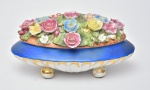 Linda caixa porta joias oval confeccionada em porcelana, em forma de floreira, apresentando riquíssima policromia, decorada com flores em relevo na parte superior, pés bolacha. Med.: 11x23x13 cm.