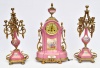 SÈVRES - Magnífica garniture em porcelana francesa na cor rosa constando um relógio e dois candelabros para cinco velas, sendo o relógio representando "Cena romântica" na galeria central, assinada E. Raubie. Guarnições e adornos em bronze. Med.: 58 cm (relógio) e 60 cm (candelabros).