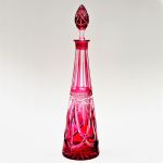 Garrafa em cristal da Bohemia com `overlay` na cor rubi, com baixo relevo em intercecção de fitas elípticas formando reservas enciamdas por raios e folhas, séc. XIX. Altura: 42 cm.