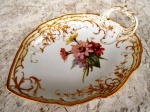 KPM - BERLIN - recipiente KONIGLICHE PORZELLAN - MANUFAKTUR - em porcelana com esmaltagem branca vitrificada  em forma de grande folha com alça , ornamentado por ouro brunido em alto relevo e ao centro pintura com ramos de flores . Peça com marca de procedência medindo 23    x 18 x 7 cm.