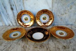 Cinco pratinhos em porcelana vitrificadas e esmaltadas , sendo quatro da marca DP com cenas   idílicas ao centro e largas faixas em ouro brunido. Um alemão com faixa azul cobalto e rendados   em ouro .Diâmetro 10/11 cm.