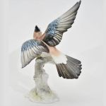Escultura de pássaro pousado com asas abertas sobre tronco em porcelana alemã HUTSCHENREUTHER, com bela pintura em marrom, azul, preto e cinza. Med: 23 x 17 cm.
